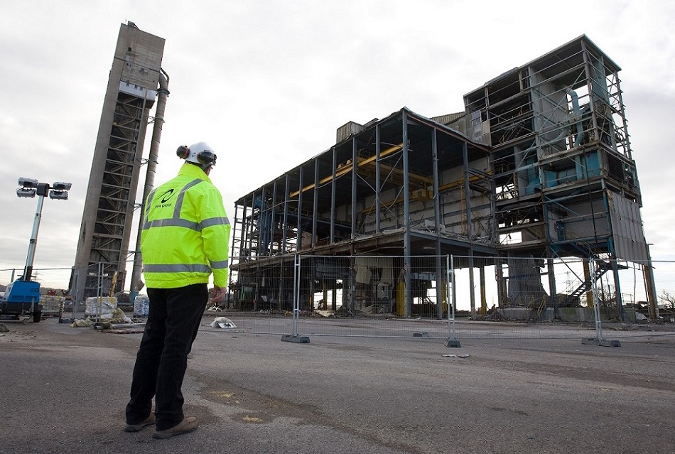 A person standing with black trousers, a neon yellow hi-visibility jacket, and white helmet on. They are looking towards an abandoned building site.